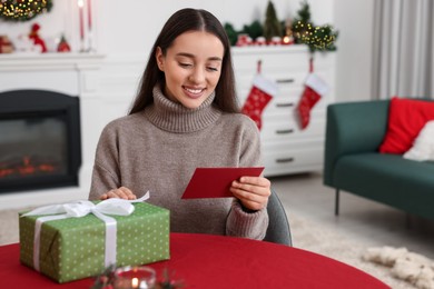 Happy young woman with Christmas gift reading greeting card at table in decorated room