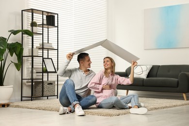 Photo of Young family housing concept. Pregnant woman with her husband sitting under cardboard roof on floor at home
