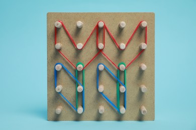 Photo of Wooden geoboard with flower shapes made of rubber bands on light blue background. Educational toy for motor skills development