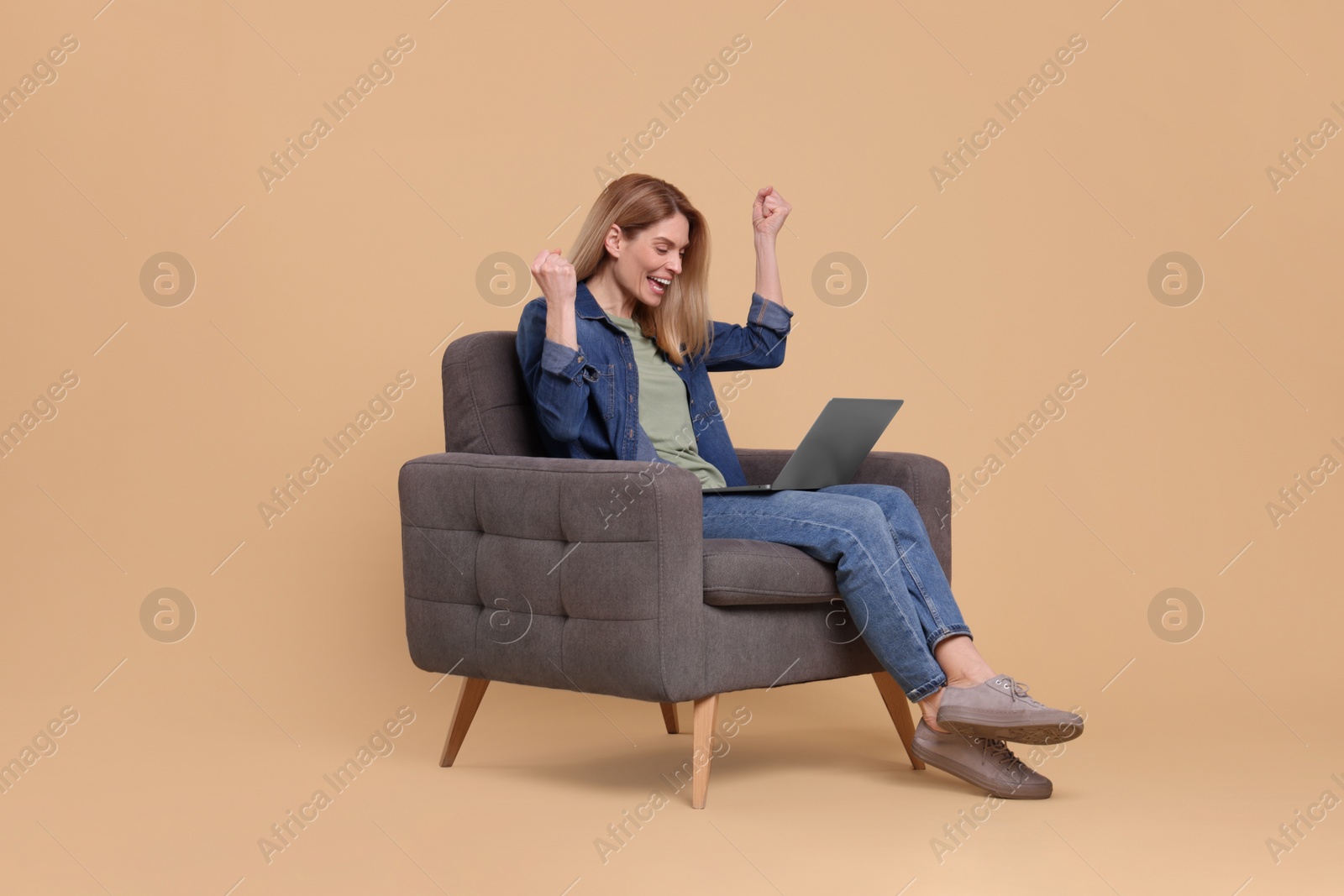 Photo of Emotional woman with laptop sitting in armchair on beige background