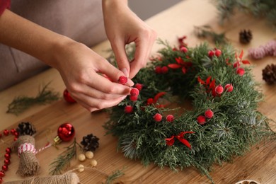 Photo of Florist making beautiful Christmas wreath with berries at wooden table indoors, closeup