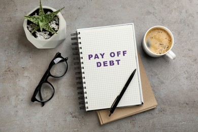 Image of Notebook with phrase Pay Off Debt, cup of aromatic coffee and plant on grey table, flat lay