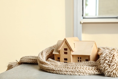 Photo of Wooden house model wrapped in scarf on grey stone table, space for text. Heating efficiency