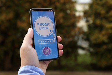 Man holding smartphone with promo code near trees outdoors, closeup. Space for text