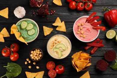 Photo of Different kinds of tasty hummus served with nachos on black wooden table, flat lay