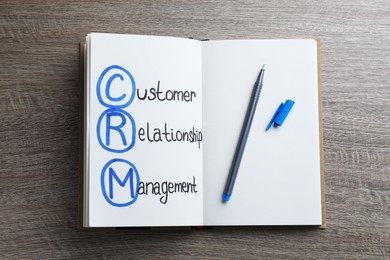 Photo of Notebook with abbreviation CRM (Customer Relationship Management) and pen on wooden background, top view