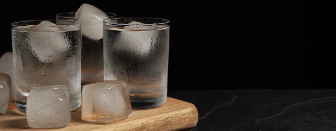 Shot glasses of vodka with ice cubes on black textured table, space for text. Banner design