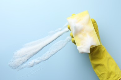 Cleaner in rubber glove holding sponge with foam on light blue background, top view