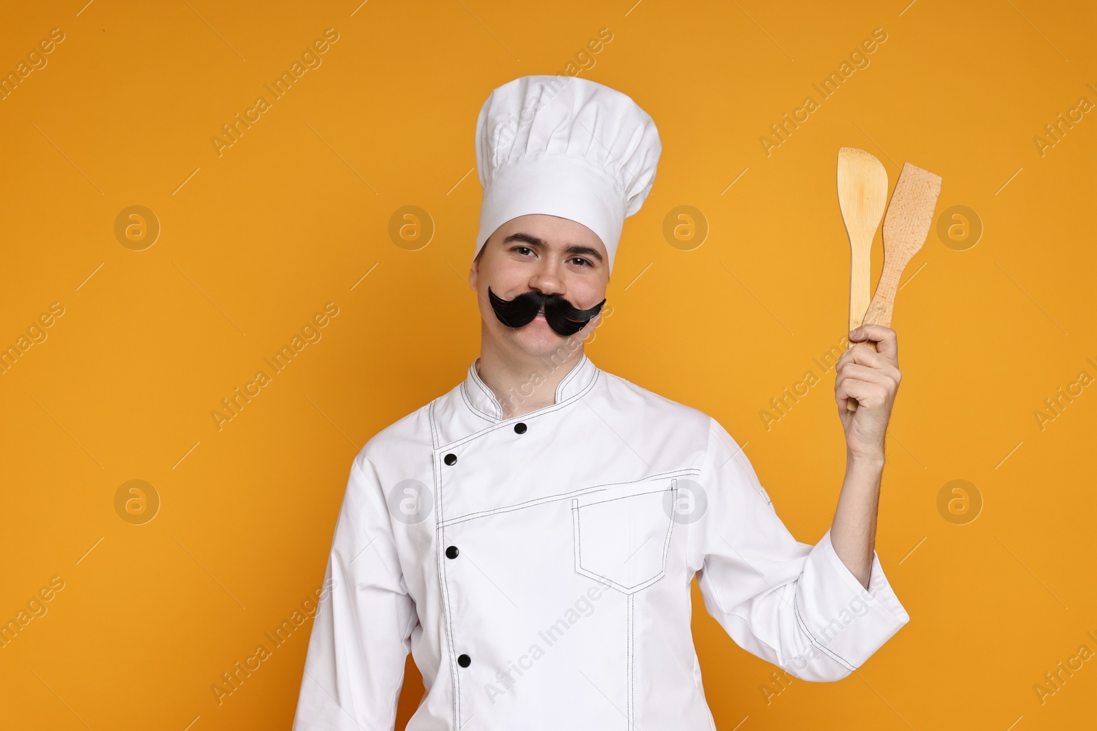 Photo of Portrait of happy confectioner with funny artificial moustache holding wooden spatulas on orange background