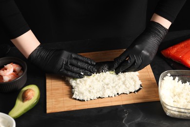 Photo of Chef in gloves wrapping sushi roll at dark textured table, closeup