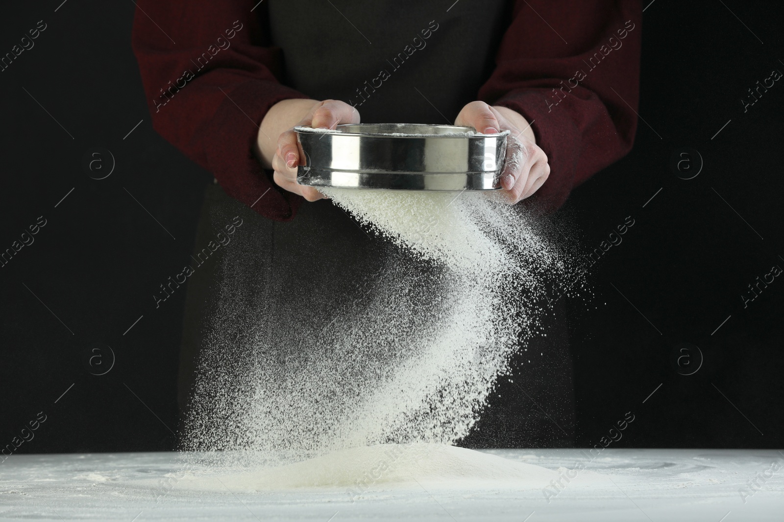 Photo of Woman sieving flour at table against black background, closeup