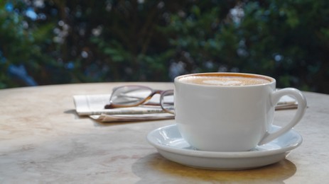 Cup of delicious coffee, glasses and newspaper on beige marble table outdoors, space for text