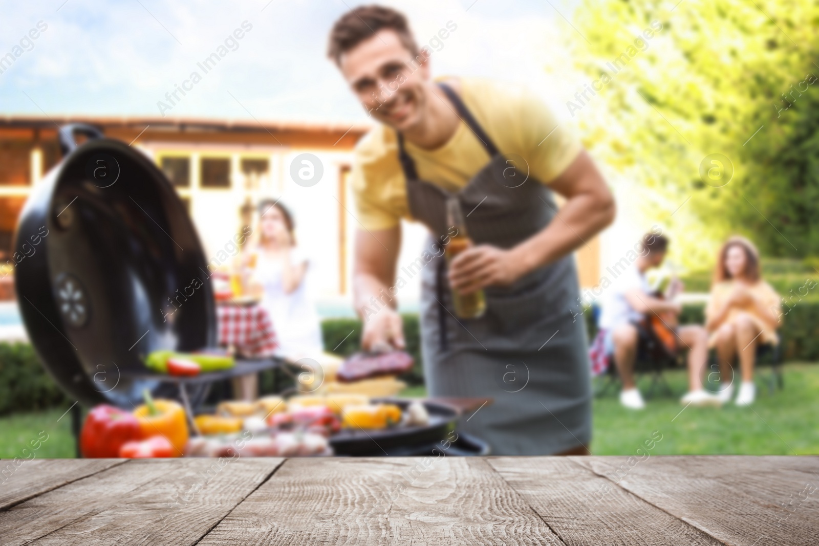 Image of Empty wooden table and blurred view of man with beer cooking on barbecue grill outdoors