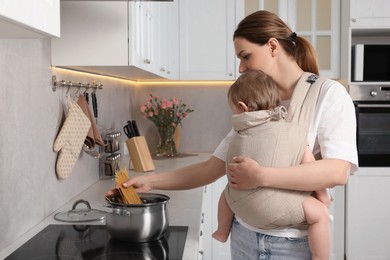 Mother holding her child in sling (baby carrier) while cooking pasta in kitchen