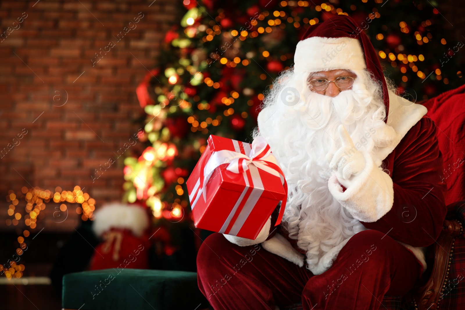 Photo of Santa Claus with gift box near Christmas tree indoors