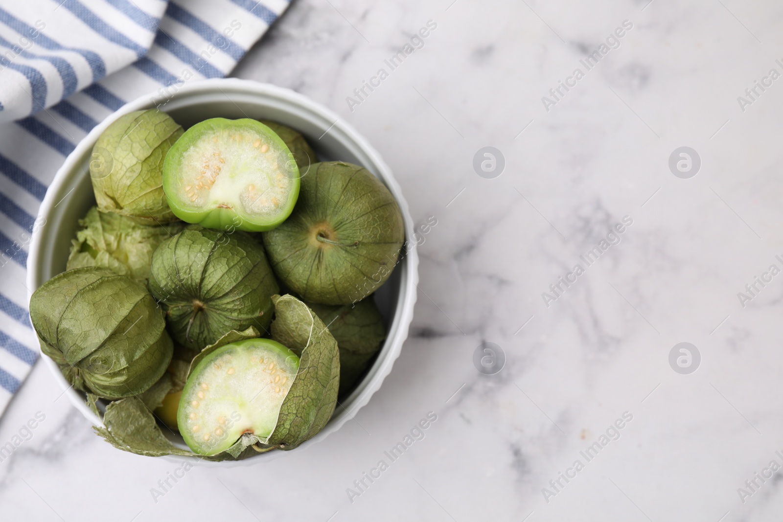 Photo of Fresh green tomatillos with husk in bowl on light marble table, top view. Space for text