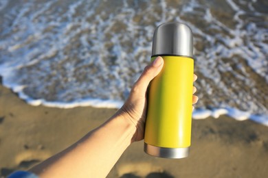 Woman holding yellow thermos with hot drink on beach near sea, closeup