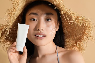Photo of Beautiful young woman in straw hat with sunscreen on her face holding sun protection cream against beige background, closeup