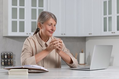 Photo of Beautiful senior woman with cup of drink near laptop at white marble table in kitchen