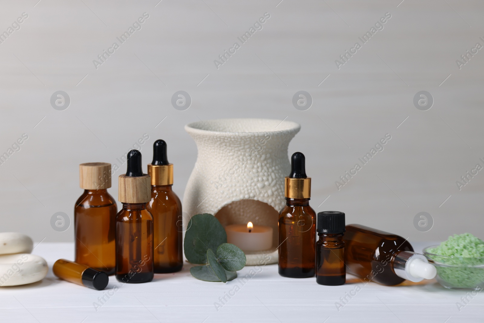 Photo of Different aromatherapy products, burning candle and eucalyptus leaves on white wooden table against light background