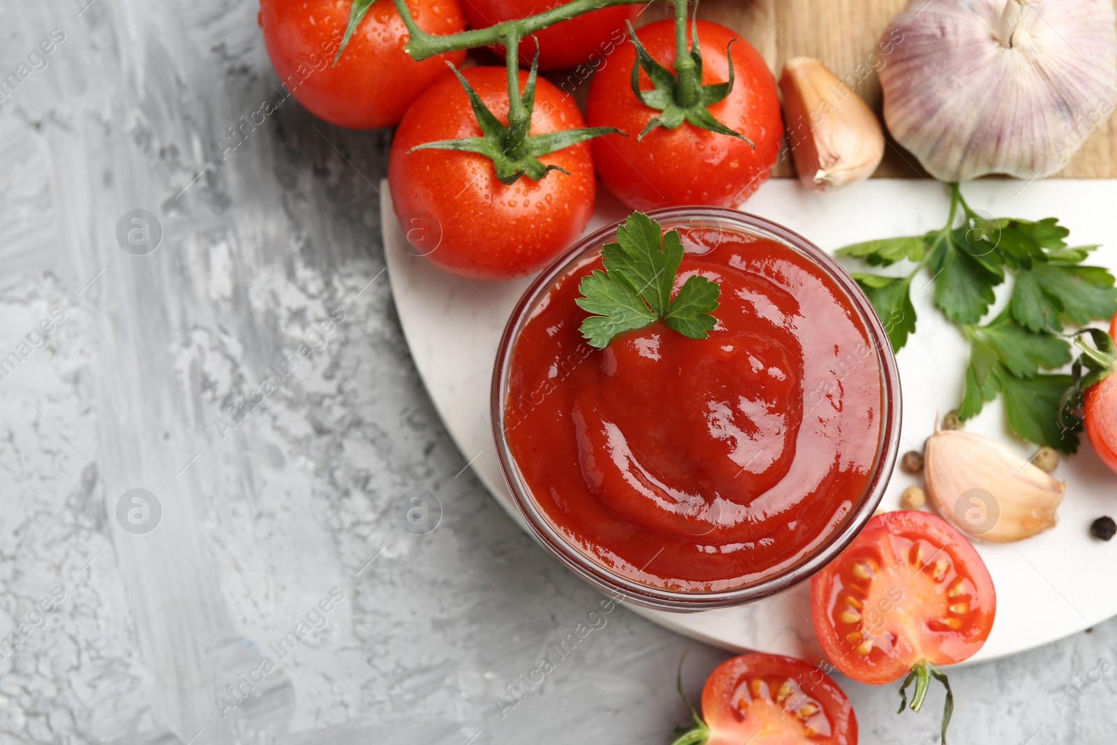 Photo of Delicious ketchup in bowl, parsley, garlic and tomatoes on grey textured table, top view. Space for text