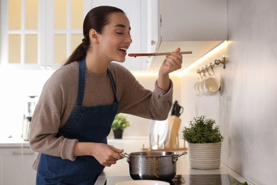 Photo of Smiling woman with wooden spoon tasting tomato soup in kitchen