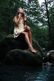 Beautiful young woman sitting on rock near mountain river in forest