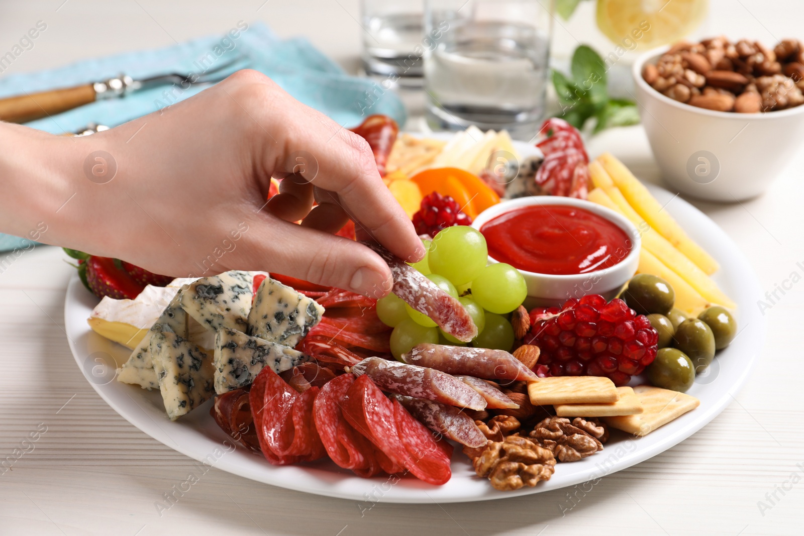 Photo of Woman taking sausage from plate with different appetizers on white wooden table, closeup