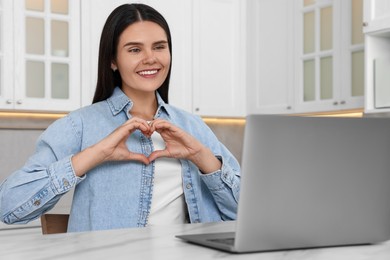 Photo of Happy young woman having video chat via laptop and making heart at table in kitchen