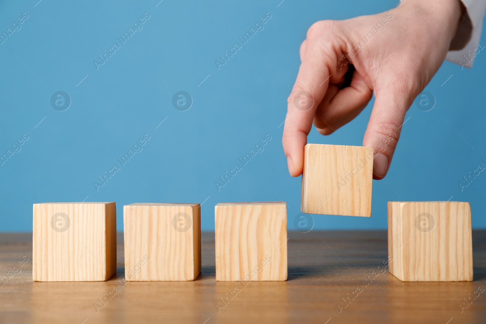 Photo of Woman arranging empty cubes in row on wooden table against light blue background, closeup. Space for text