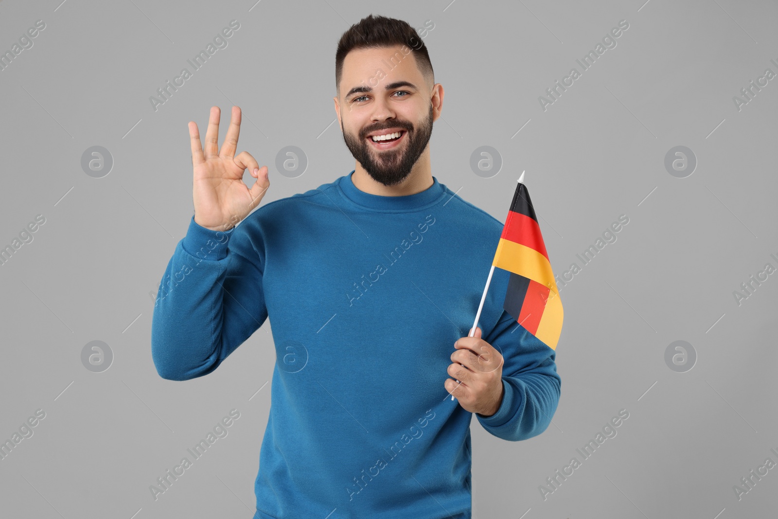 Photo of Young man with flag of Germany showing ok gesture on light grey background