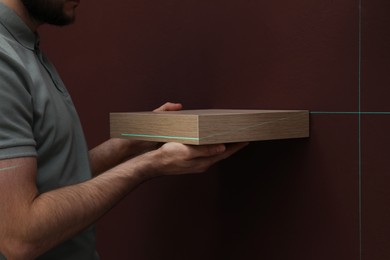 Photo of Man using cross line laser level for hanging shelf on brown wall, closeup