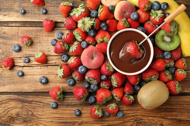 Photo of Fondue fork with strawberry in bowl of melted chocolate surrounded by other fruits on wooden table, flat lay