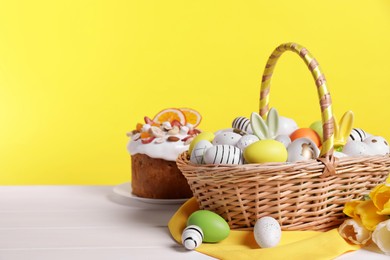 Photo of Wicker basket with festively decorated Easter eggs and beautiful tulips on white wooden table against yellow background. Space for text