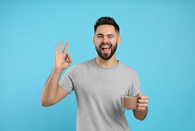 Photo of Young man with glass cup of chocolate milk showing Ok on light blue background
