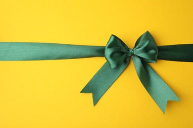 Green satin ribbon with bow on yellow background, top view