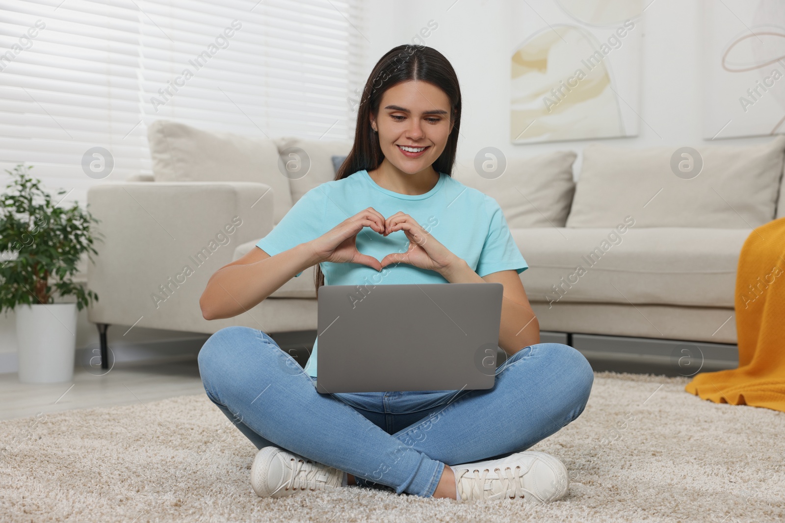 Photo of Happy young woman having video chat via laptop and making heart on floor in living room