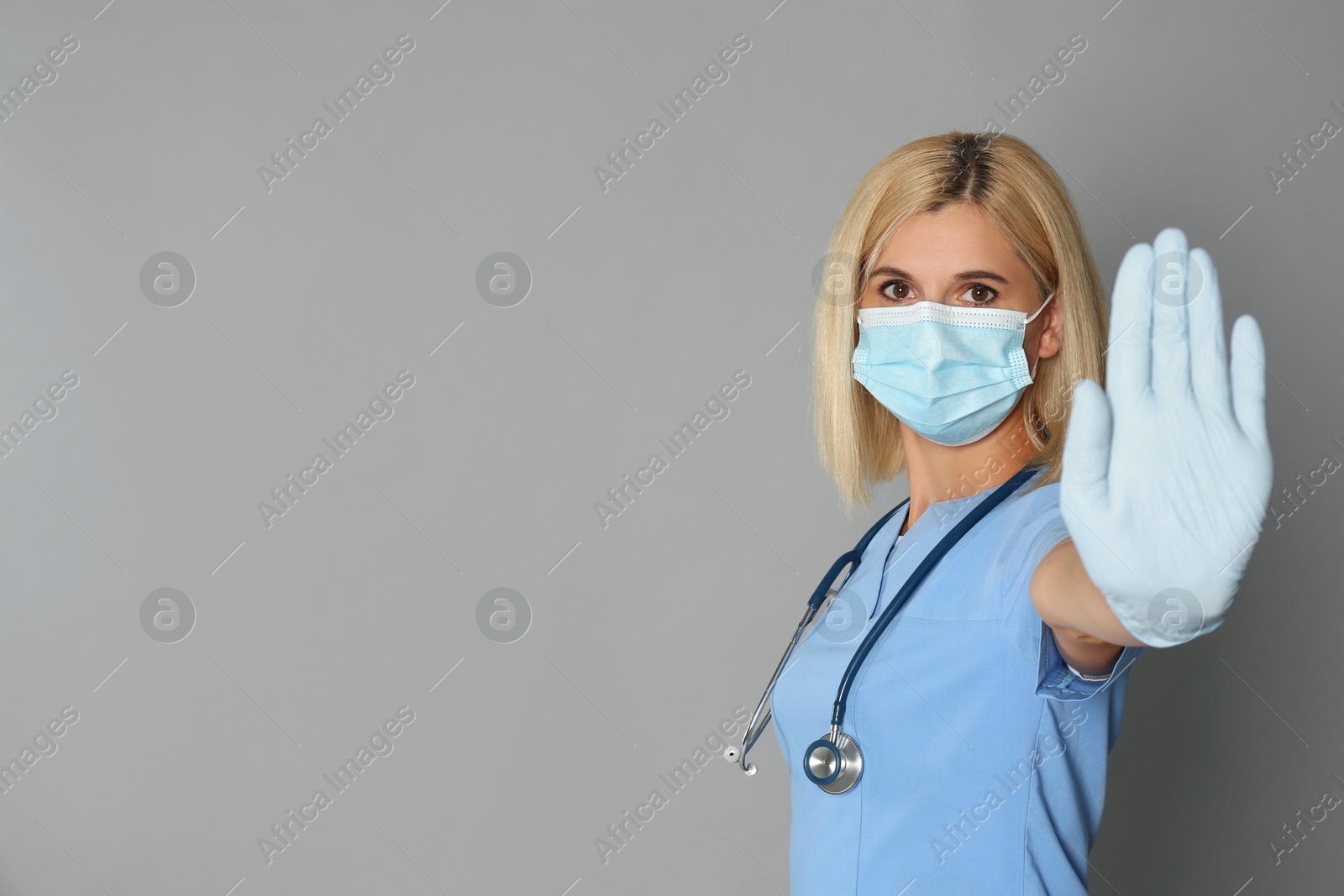 Photo of Doctor in protective mask showing stop gesture on grey background, space for text. Prevent spreading of coronavirus