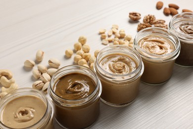 Many tasty nut butters in jars and nuts on white wooden table, closeup