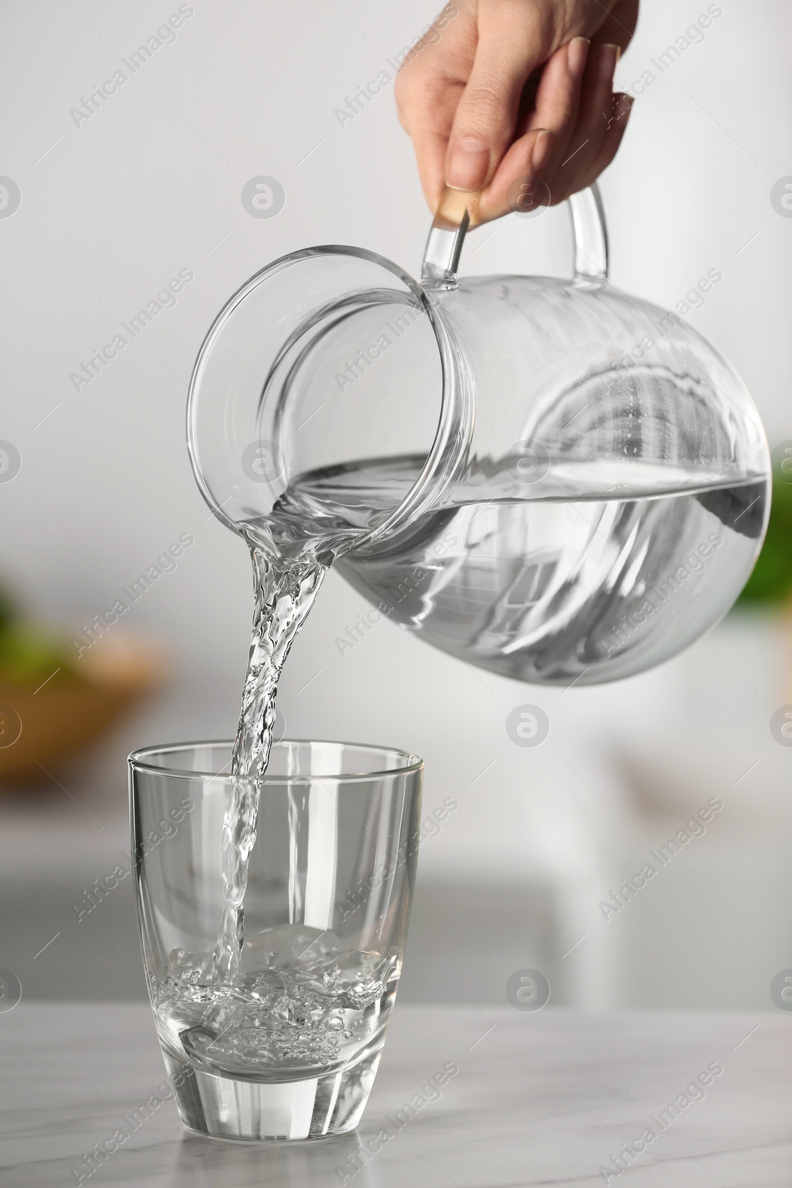 Photo of Woman pouring water from jug into glass on white marble table in kitchen, closeup