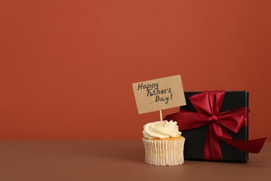 Photo of Happy Father's Day. Tasty cupcake with greetings and gift box on brown table. Space for text