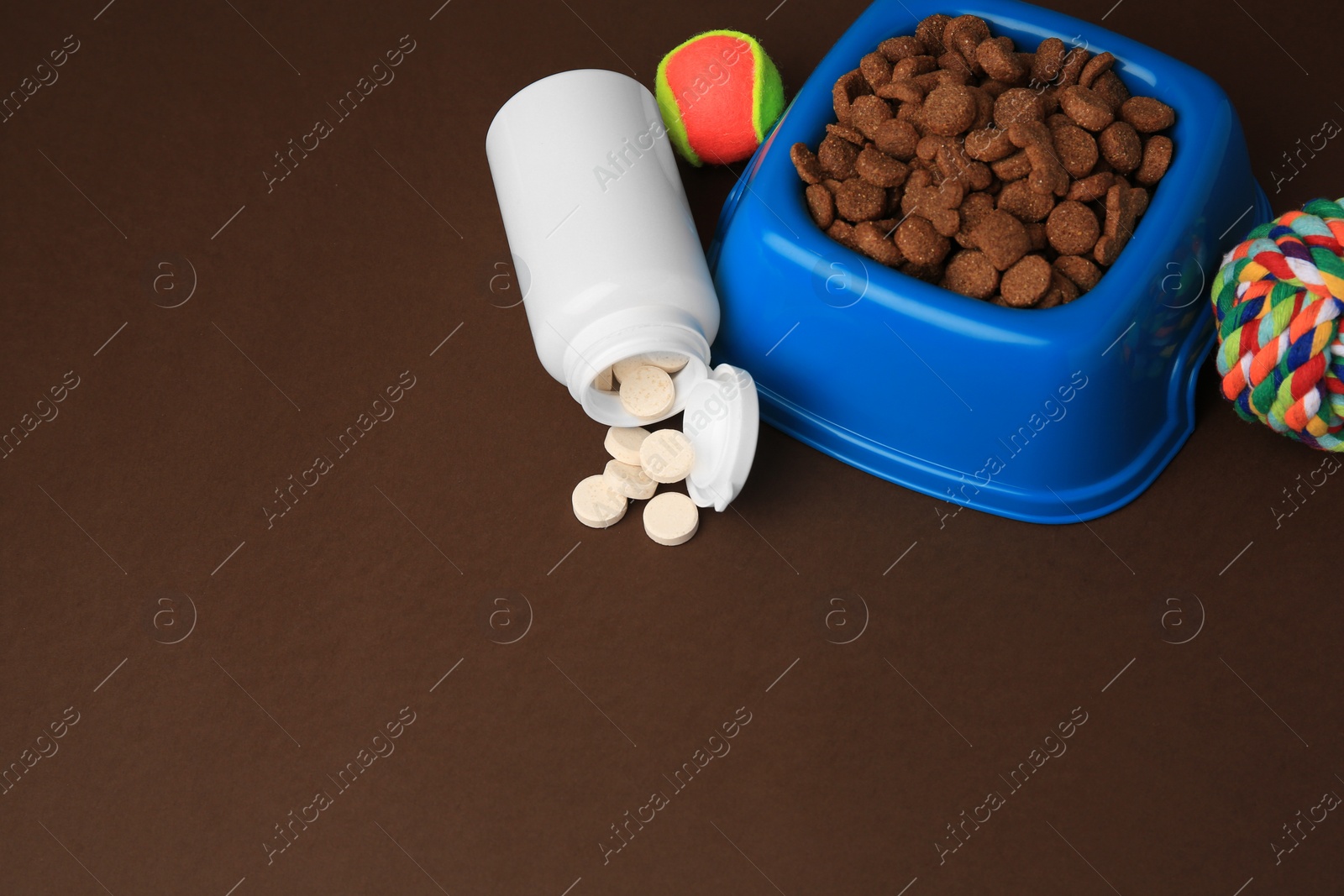 Photo of Bowl with dry pet food, bottle of vitamins and toys on brown background. Space for text