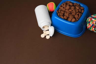 Bowl with dry pet food, bottle of vitamins and toys on brown background. Space for text