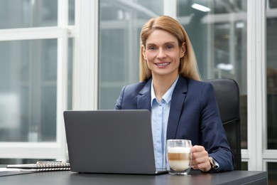 Photo of Woman with cup of coffee working on laptop at black desk in office