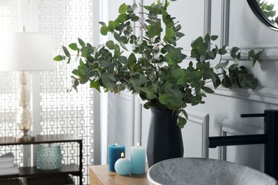 Photo of Eucalyptus branches and burning candles near vessel sink on bathroom vanity. Interior design