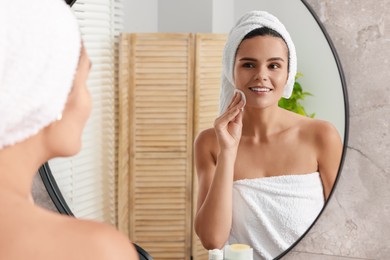 Young woman cleaning her face with cotton pad near mirror in bathroom