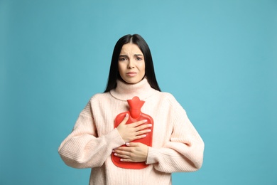 Woman using hot water bottle to relieve chest pain on light blue background