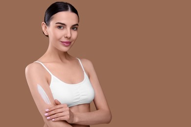 Beautiful woman with smear of body cream on her arm against light brown background, space for text