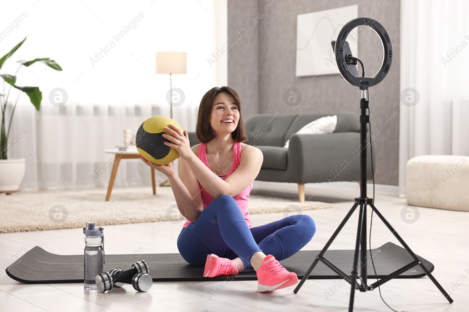Photo of Happy sports blogger holding medicine ball while streaming online fitness lesson with smartphone at home