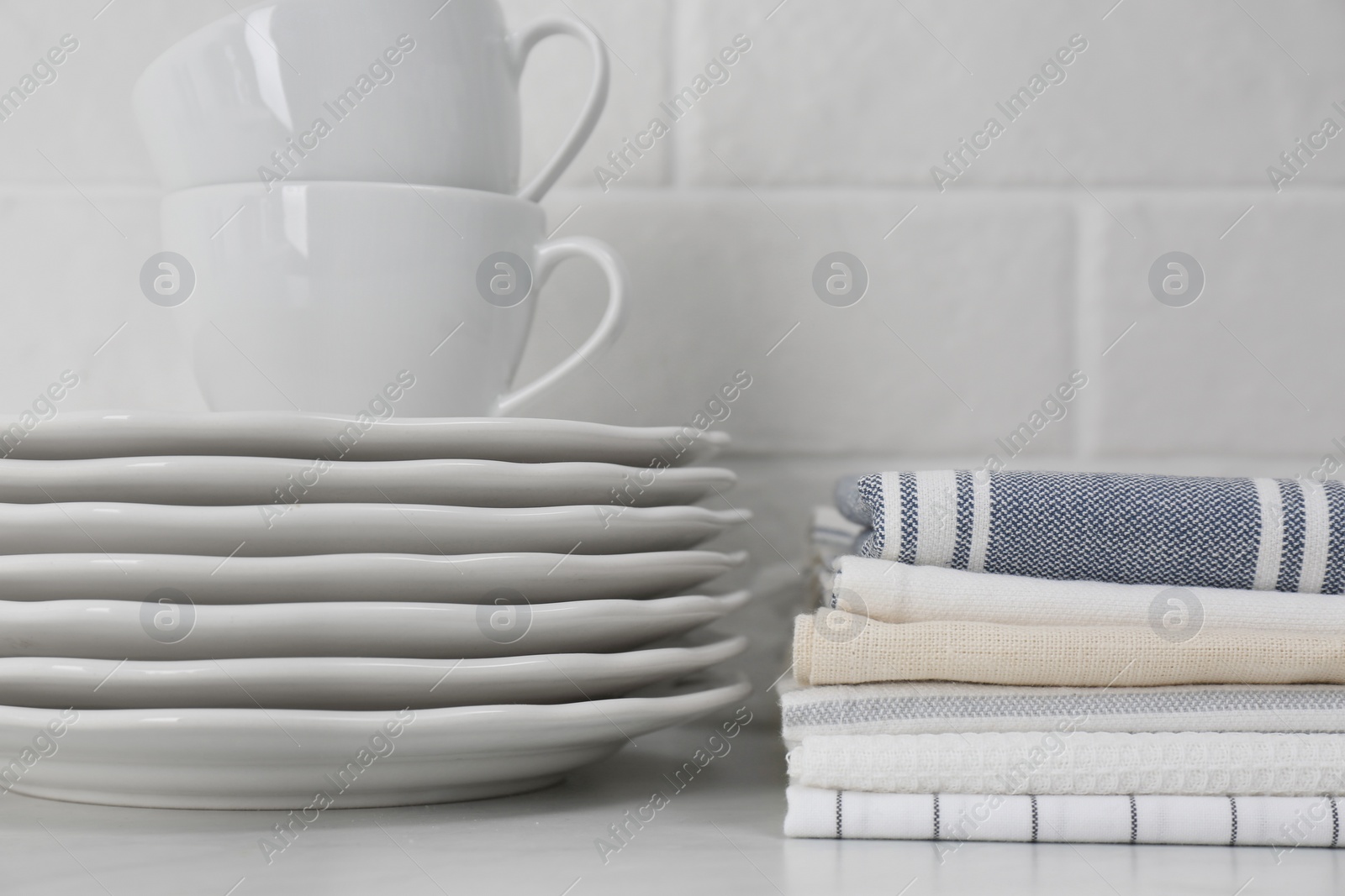 Photo of Stack of soft kitchen towels and dishware on table near white brick wall, closeup